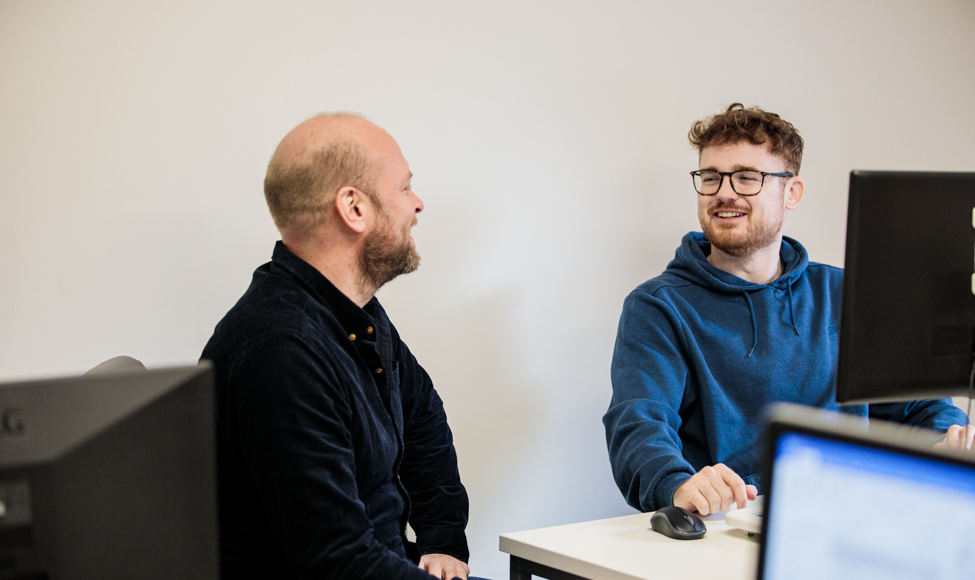 2 men smiling sat at a desk on a computer